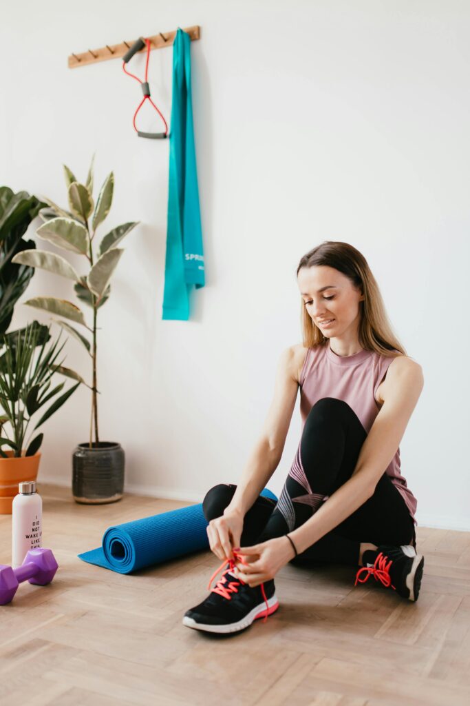 pexels-photo-4498551-4498551 Young woman tying shoelaces in a home gym setting with fitness equipment.