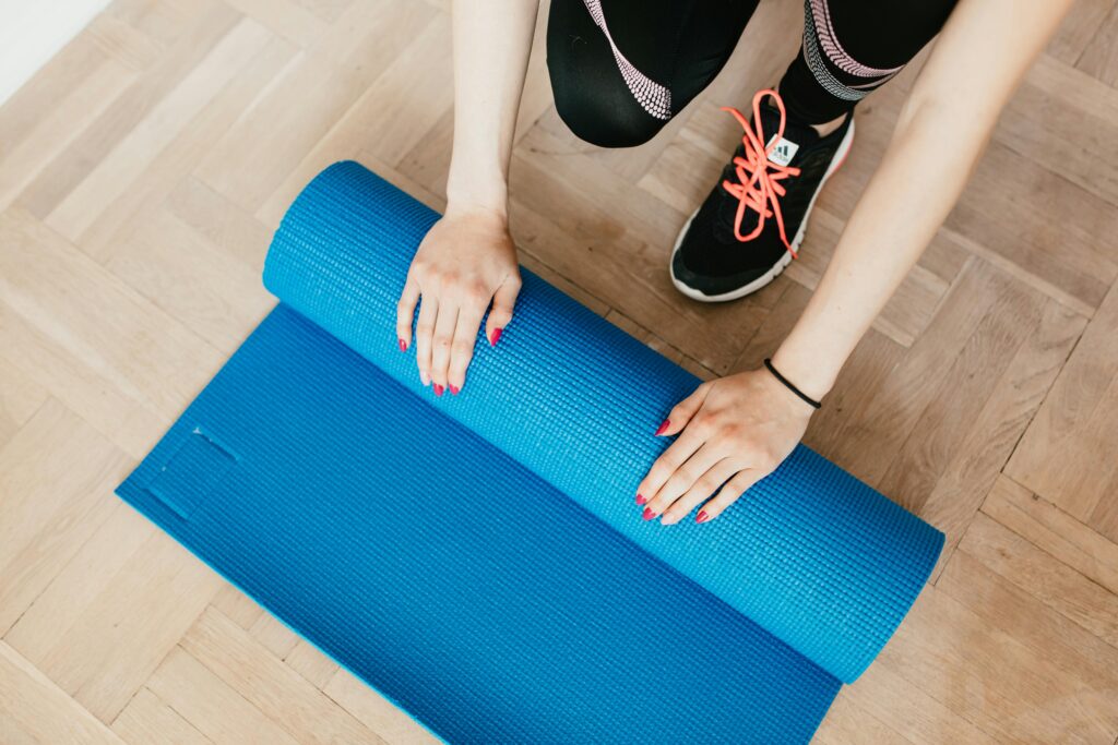 From above of unrecognizable female athlete in leggings and sneakers unfolding sport mat for practicing yoga in morning at home