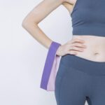 Crop unrecognizable slim female wearing purple sportswear standing with hand on waist holding resistance bands against white background