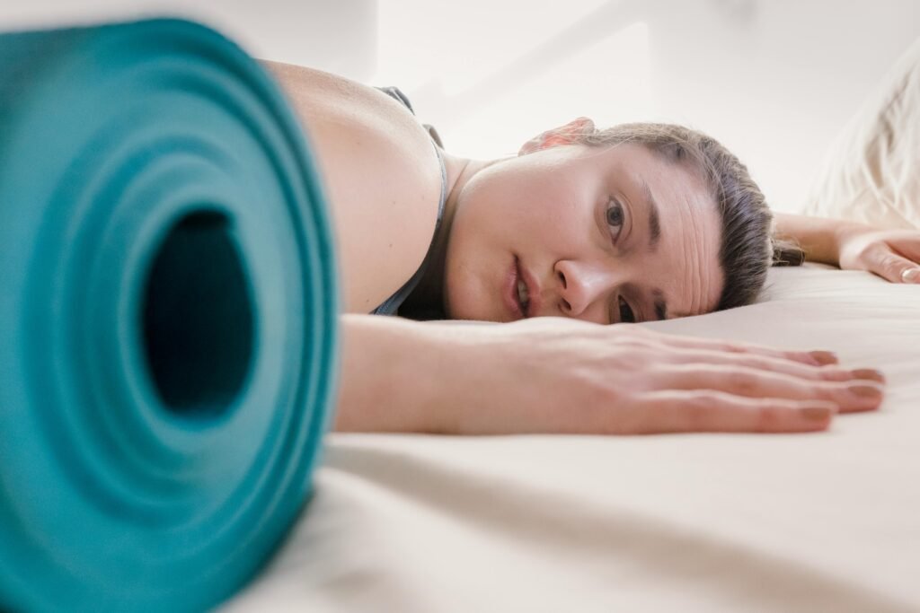 pexels-photo-6697279-6697279 Exhausted female looking at camera while lying on comfortable bed with blanket and rolled mat during training in light room
